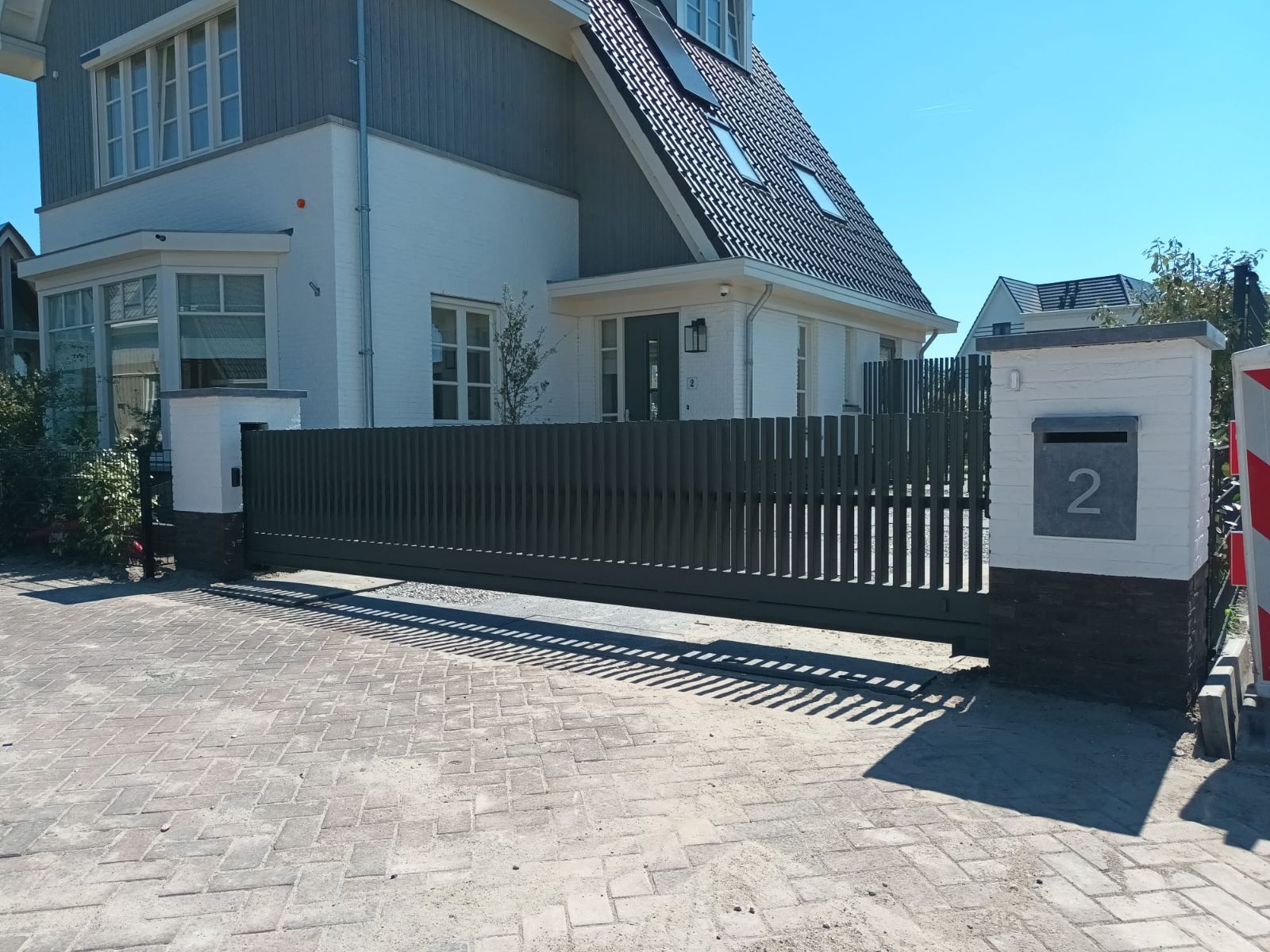Stalen schuifpoort naast een witte poortpilaar met brievenbus en huisnummer